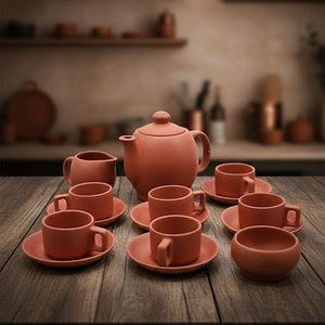 May include: A terracotta-colored tea set arranged on a wooden table. The set includes a teapot, cups, saucers, a creamer, and a bowl. The background features a blurred kitchen setting with shelves and other items.