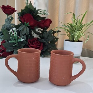 Puede incluir: Dos tazas de terracota con textura de ladrillo y asas curvas. Están sobre una superficie blanca, con rosas rojas y vegetación de fondo, y una planta en maceta. Las tazas son de terracota.