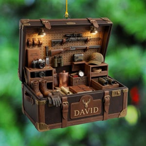 May include: A brown trunk-shaped ornament featuring the name DAVID and a deer emblem. The interior is a pegboard display containing a rifle, shotgun, binoculars, cameras, a leather field guide, a flask, boots, gloves, and a cap with two crossed rifles.
