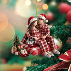 May include: A hanging Christmas ornament featuring a couple in matching red and white plaid pyjamas and Santa hats labelled Olive and Daniel. They sit on a plaid cushion with a small ultrasound image, a miniature Christmas tree, and gifts.