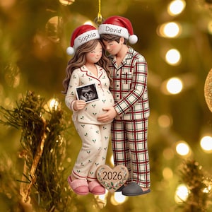 May include: A hanging Christmas ornament featuring a pregnant person and a partner in matching pajamas. The person wears cream pajamas with holly, and the partner wears plaid pajamas. Both wear Santa hats labeled Sophia and David, holding an ultrasound image. A heart at the base displays the year 2026.
