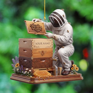 Op de afbeelding: Een decoratief ornament met een imker in een wit beschermend pak die naast een houten bijenkorf met drie niveaus knielt. De tekst YOUR NAME staat op het bovenste gedeelte. Bijen, zonnebloemen en lavendel omringen de basis van het ornament.