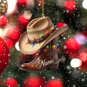 May include: A hanging ornament featuring a tan straw cowboy hat with a braided band and a brown leather cowboy boot with a silver spur. The items are wrapped in colorful string lights, and the word Name is written in white script on the boot.