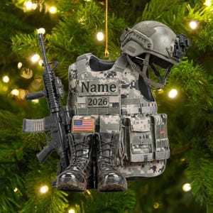 Könnte beinhalten: Ein hängendes Ornament mit einer taktischen Weste in digitalem Tarnmuster mit dem Text Name und 2026, einem schwarzen Gewehr, einem Paar schwarzer Kampfstiefel, einem kleinen US-Flaggenaufnäher und einem taktischen Helm, befestigt an einer goldenen Schnur.