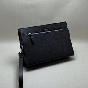 Könnte beinhalten: Eine schwarze Leder-Clutch mit strukturierter Oberfläche. Die Clutch hat einen silbernen Reißverschluss und eine Handschlaufe. Das Design ist schlicht und elegant, passend für verschiedene Anlässe.