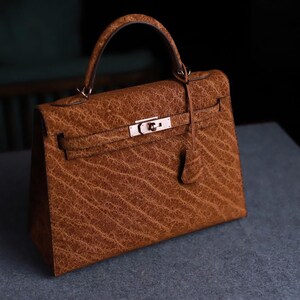 Könnte beinhalten: Eine braune Lederhandtasche mit einem oberen Griff und einem silberfarbenen Verschluss. Die Tasche hat eine strukturierte Oberfläche und einen Klappenverschluss. Die Handtasche steht auf einer grauen Oberfläche.