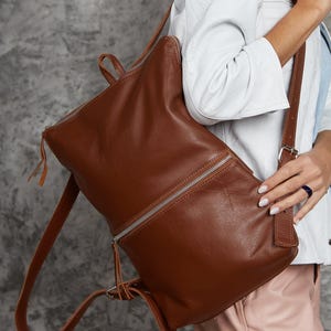 Peut inclure: Un sac à dos en cuir marron avec une fermeture éclair argentée. Le sac à dos a des bretelles réglables et une poignée supérieure. Le sac est en cuir lisse et texturé, de conception minimaliste.