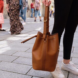 Könnte beinhalten: Eine braune Leder-Beuteltasche mit Kordelzug und Schulterriemen. Die Tasche hat goldfarbene Beschläge. Sie wird von einer Person getragen, die eine schwarze Hose und weiße Turnschuhe trägt. Im Hintergrund eine belebte Straße.