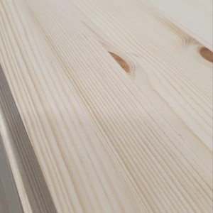 May include: Close-up of stacked, unfinished pine wood planks. The wood grain is visible, with knots and a light, natural colour. The planks are ready for construction or woodworking projects.