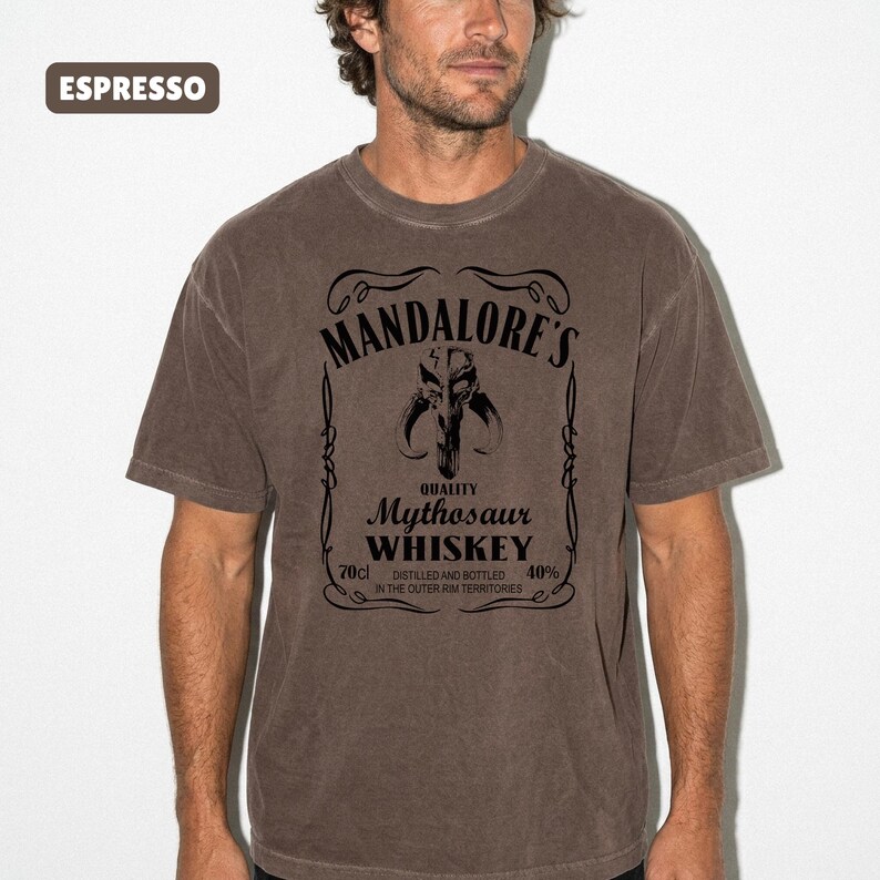 Puede incluir: Camiseta color espresso con un dise&ntilde;o gr&aacute;fico negro. El dise&ntilde;o incluye el texto "MANDALORE'S" encima de una calavera de Mythosaur, con "WHISKEY" debajo. La camiseta tambi&eacute;n muestra "70cl" y "40%" con texto adicional. Cuello redondo.