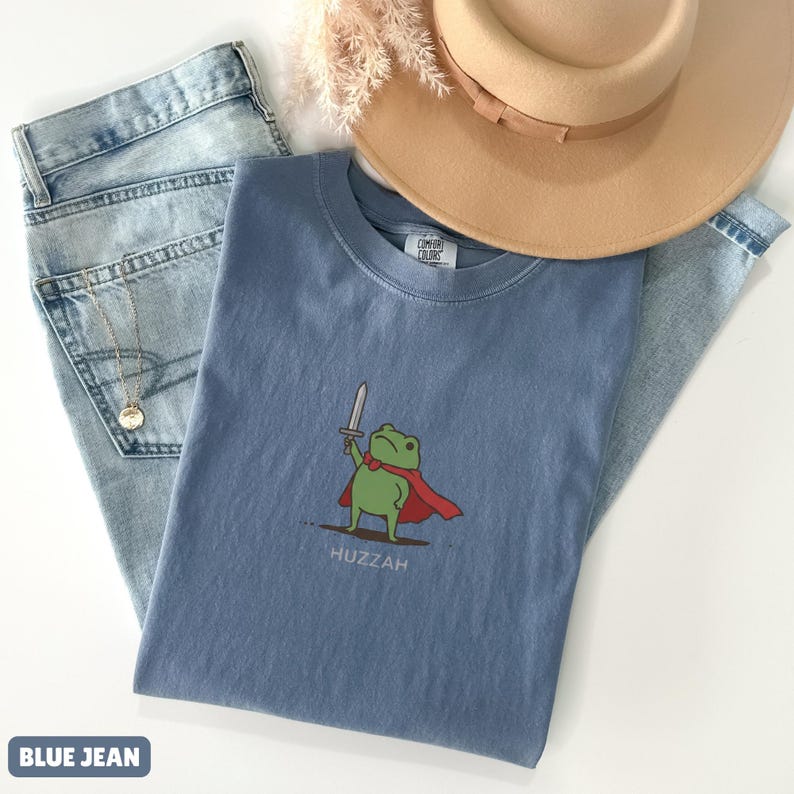 May include: A light blue t-shirt featuring a cartoon frog with a sword and cape, with the word "Huzzah" below. The shirt is paired with light wash blue jeans and a tan hat.