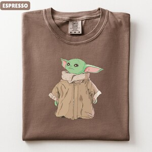 May include: Espresso-colored t-shirt featuring a graphic of a green alien character wearing a tan coat. The shirt has a crew neck and a tag that reads "Comfort Colors". The folded shirt is a medium size.