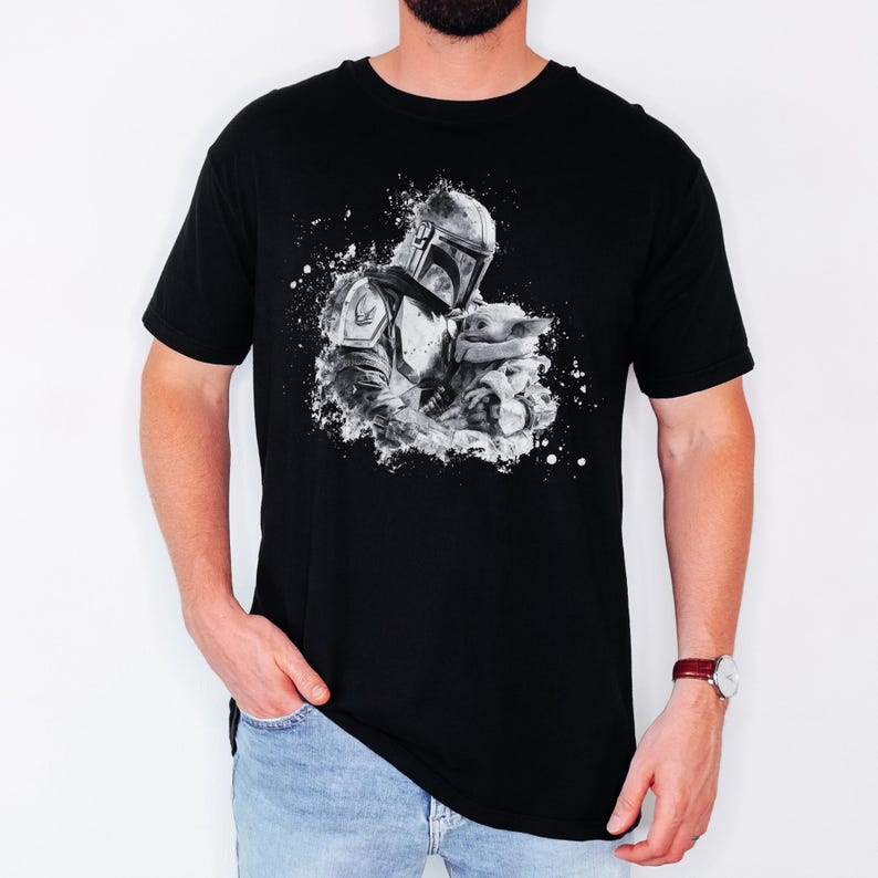 Op de afbeelding: Zwart T-shirt met een zwart-witte afbeelding van de Mandalorian en Grogu. Het ontwerp heeft een spatteffect, met de figuren in het midden. Het shirt heeft korte mouwen en een ronde hals.