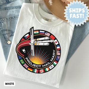 Könnte beinhalten: Weißes T-Shirt mit einem kreisförmigen Grafikdesign, das ein Raumschiff, den Text "PROJECT HAIL MARY" und Flaggen verschiedener Nationen zeigt. Der Text "WHITE" ist ebenfalls unten zu sehen.