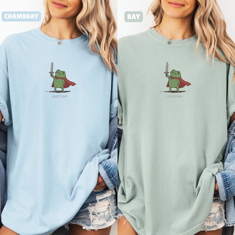 May include: Light blue and light green t-shirts featuring a cartoon frog character with a sword and cape. The frog is green with a red cape and holds a silver sword. The word "Huzzah" is printed below the frog.