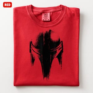 May include: A folded red t-shirt with a black helmet graphic. The shirt has a crew neck and a tag that reads "Comfort Colors". The word "RED" is in a red box in the upper left corner.