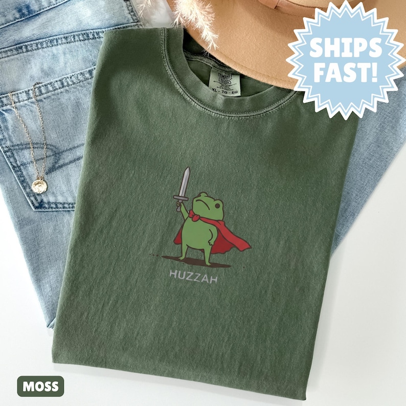 May include: A moss green t-shirt featuring a cartoon frog wearing a red cape and holding a sword. The word "Huzzah" is printed below the frog. The shirt is displayed on top of blue jeans and a tan hat.