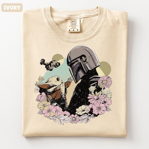 May include: Ivory-colored t-shirt featuring a graphic of a helmeted figure and a small creature surrounded by flowers. The design includes a spaceship and butterflies. The shirt has a crew neck and a tag that reads "Comfort Colors".