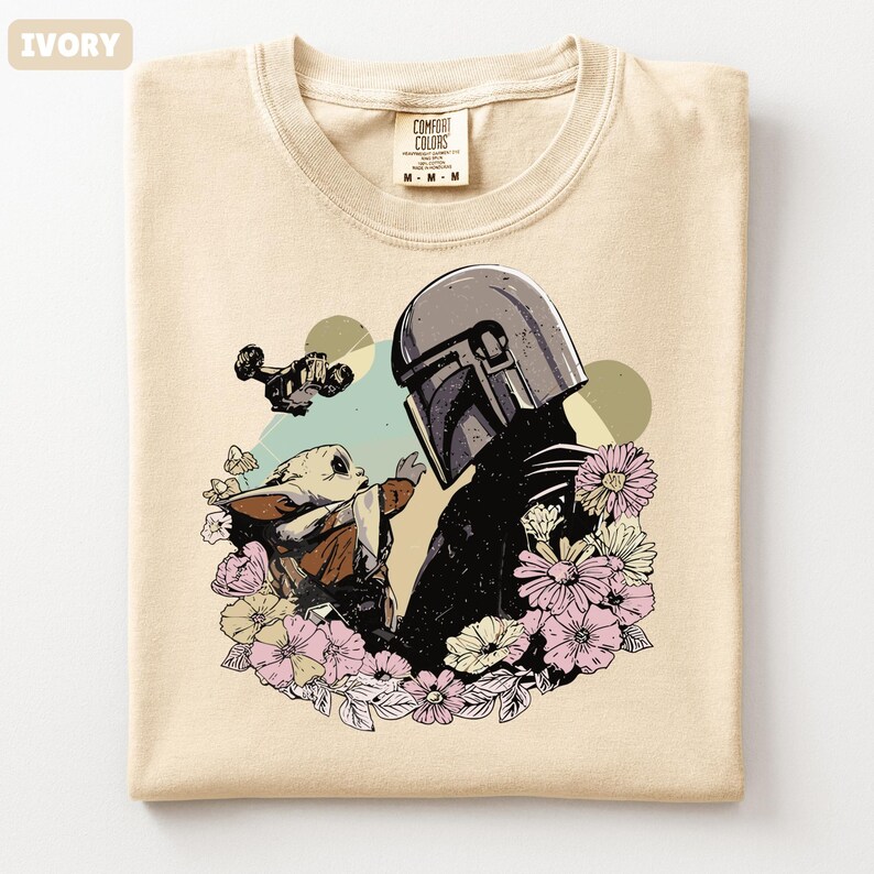 May include: Ivory-colored t-shirt featuring a graphic of a helmeted figure and a small creature surrounded by flowers. The design includes a spaceship and butterflies. The shirt has a crew neck and a tag that reads "Comfort Colors".