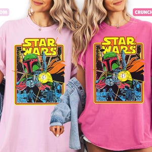 May include: Two t-shirts, one light pink and one fuchsia, each featuring a vintage Star Wars graphic. The graphic includes the words "STAR WARS" in yellow, and an illustration of Boba Fett. The shirts have short sleeves.