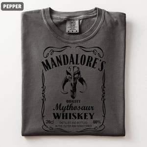 Puede incluir: Camiseta color pimienta con un gr&aacute;fico negro. El gr&aacute;fico dice "MANDALORE'S Mythosaur WHISKEY" con una imagen de calavera de Mythosaur. El texto incluye "70cl" y "40%" y "DESTILADO Y EMBOTELLADO EN LOS TERRITORIOS DEL BORDE EXTERIOR".
