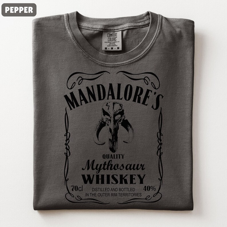 Puede incluir: Camiseta color pimienta con un gr&aacute;fico negro. El gr&aacute;fico dice "MANDALORE'S Mythosaur WHISKEY" con una imagen de calavera de Mythosaur. El texto incluye "70cl" y "40%" y "DESTILADO Y EMBOTELLADO EN LOS TERRITORIOS DEL BORDE EXTERIOR".