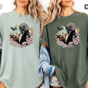 May include: Two t-shirts, one in a light green "Bay" color and the other in a dark green "Moss" color. Each shirt features a graphic of a character in a helmet with a baby character surrounded by flowers. The shirts are made of a soft material.