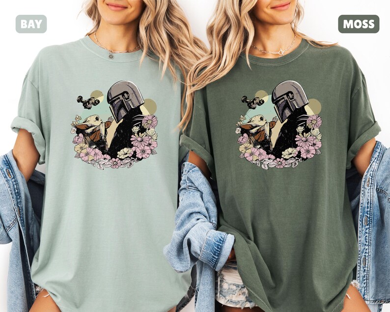 May include: Two t-shirts, one in a light green "Bay" color and the other in a dark green "Moss" color. Each shirt features a graphic of a character in a helmet with a baby character surrounded by flowers. The shirts are made of a soft material.