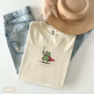 May include: Ivory-colored t-shirt featuring a cartoon frog with a sword and cape, with the word "Huzzah" below. The shirt is paired with light blue jeans, a tan hat, and a necklace.