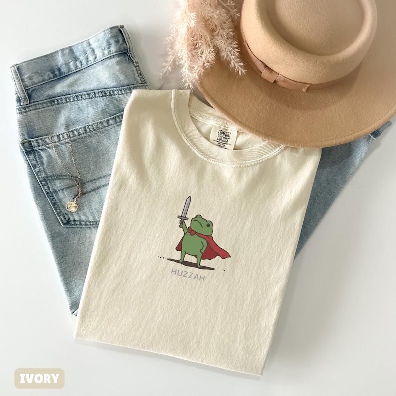 May include: Ivory-colored t-shirt featuring a cartoon frog with a sword and cape, with the word "Huzzah" below. The shirt is paired with light blue jeans, a tan hat, and a necklace.