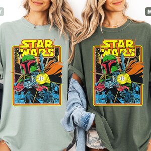 May include: Two t-shirts, one in bay and one in moss, each featuring a Star Wars graphic. The graphic includes the text "STAR WARS" in yellow, and an illustration of Boba Fett. The shirts are short-sleeved.