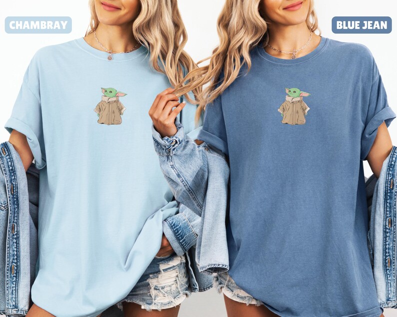 May include: Two t-shirts, one light blue and one blue, each featuring an embroidered image of a green alien character in a tan coat. The light blue shirt has the word "CHAMBRAY" printed on it. The blue shirt has the words "BLUE JEAN" printed on it.