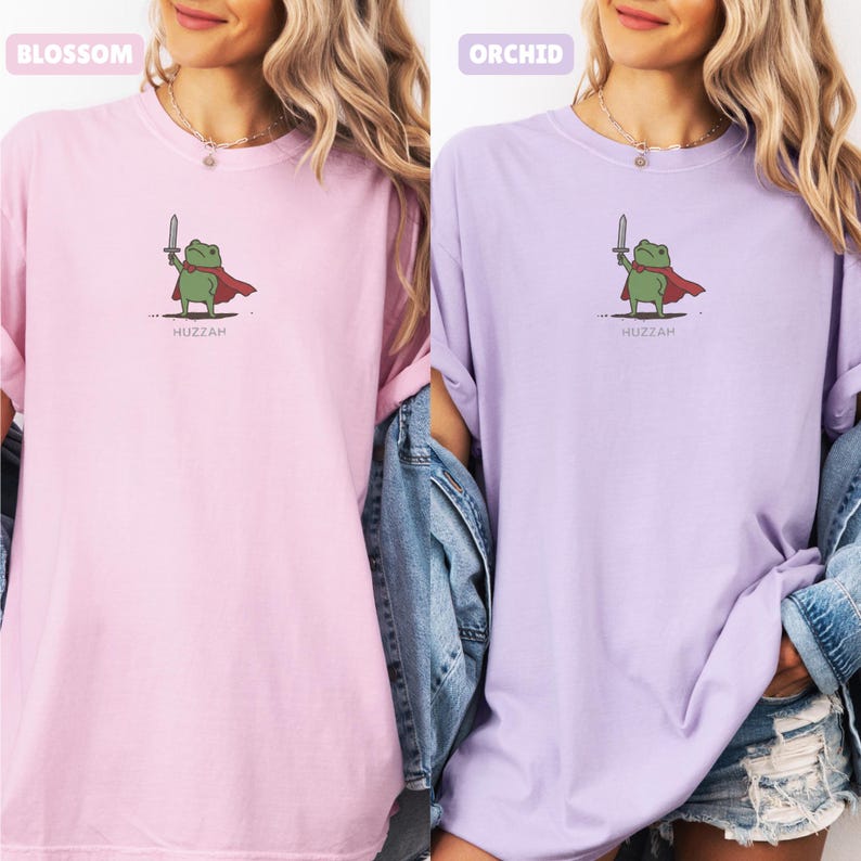 May include: Two t-shirts, one light pink and one lavender, featuring a cartoon frog with a sword and cape. The word "Huzzah" is printed below the frog. The shirt colors are labeled "Blossom" and "Orchid".