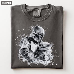 Op de afbeelding: Pepperkleurig T-shirt met een zwart-witte afbeelding van een Mandalorian figuur die Grogu vasthoudt. Het ontwerp heeft een distressed, splatter-paint effect. Het shirt heeft een ronde hals en een Comfort Colors label.