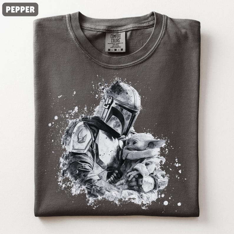 Op de afbeelding: Pepperkleurig T-shirt met een zwart-witte afbeelding van een Mandalorian figuur die Grogu vasthoudt. Het ontwerp heeft een distressed, splatter-paint effect. Het shirt heeft een ronde hals en een Comfort Colors label.