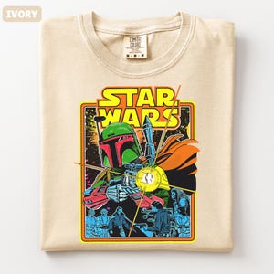 May include: Ivory-colored t-shirt featuring a Star Wars graphic. The design includes the text "STAR WARS" in yellow, with an image of Boba Fett and other characters. The shirt is a Comfort Colors brand, size M-M-M.