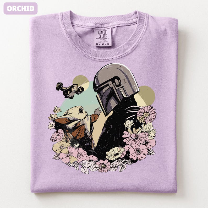 May include: Orchid-colored t-shirt featuring a graphic of a helmeted figure and a small creature surrounded by flowers. The design includes a spaceship and two yellow circles. The shirt is made of Comfort Colors material.