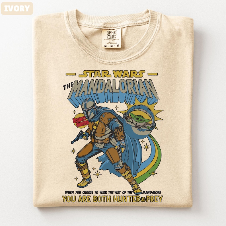 以下が含まれることがあります： スターウォーズのマンダロリアンが描かれたアイボリー色のTシャツ。デザインにはマンダロリアンのキャラクター、グローグー、そして「マンダロリアン」と「あなたがマンダロアの道を歩むことを選ぶとき、あなたはハンターと獲物の両方である」というテキストが含まれています。