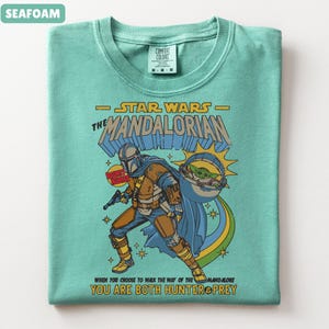 以下が含まれることがあります： スターウォーズのマンダロリアンをフィーチャーした、シーフォームグリーンのTシャツ。デザインにはマンダロリアン、グローグー、そして「When you choose to walk the way of the Mandalore, you are both hunter & prey.」というテキストが含まれています。