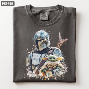 Puede incluir: Camiseta color pimienta con una ilustraci&oacute;n estilo acuarela de un personaje mandaloriano y Grogu. El dise&ntilde;o incluye un casco, armadura y Grogu envuelto en una manta. La camiseta tiene cuello redondo y una etiqueta Comfort Colors.