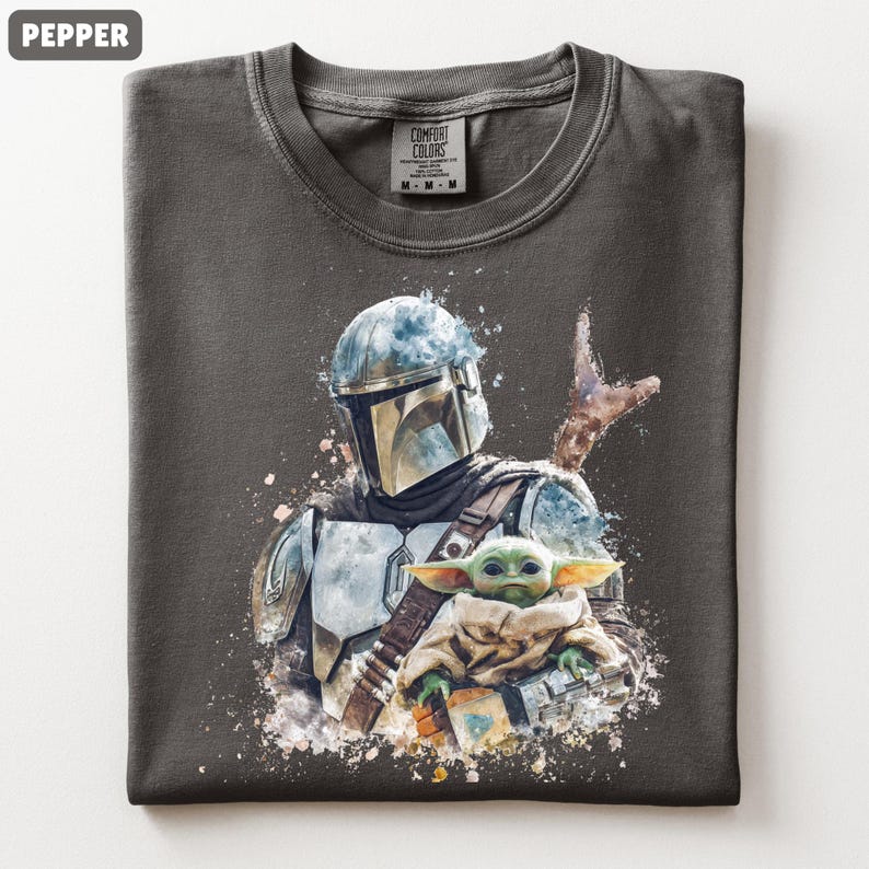 Puede incluir: Camiseta color pimienta con una ilustraci&oacute;n estilo acuarela de un personaje mandaloriano y Grogu. El dise&ntilde;o incluye un casco, armadura y Grogu envuelto en una manta. La camiseta tiene cuello redondo y una etiqueta Comfort Colors.