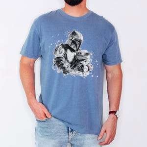 Op de afbeelding: Lichtblauw t-shirt met een zwart-witte afbeelding van een Mandalorian karakter en een klein groen wezen. De afbeelding heeft een spat effect. Het shirt heeft korte mouwen en een ronde hals.