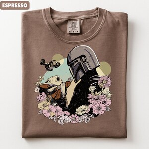 May include: Espresso-colored t-shirt featuring a graphic of a helmeted figure and a small creature surrounded by flowers. The design includes a spaceship and a light green and yellow background. The shirt has a crew neck and a Comfort Colors tag.