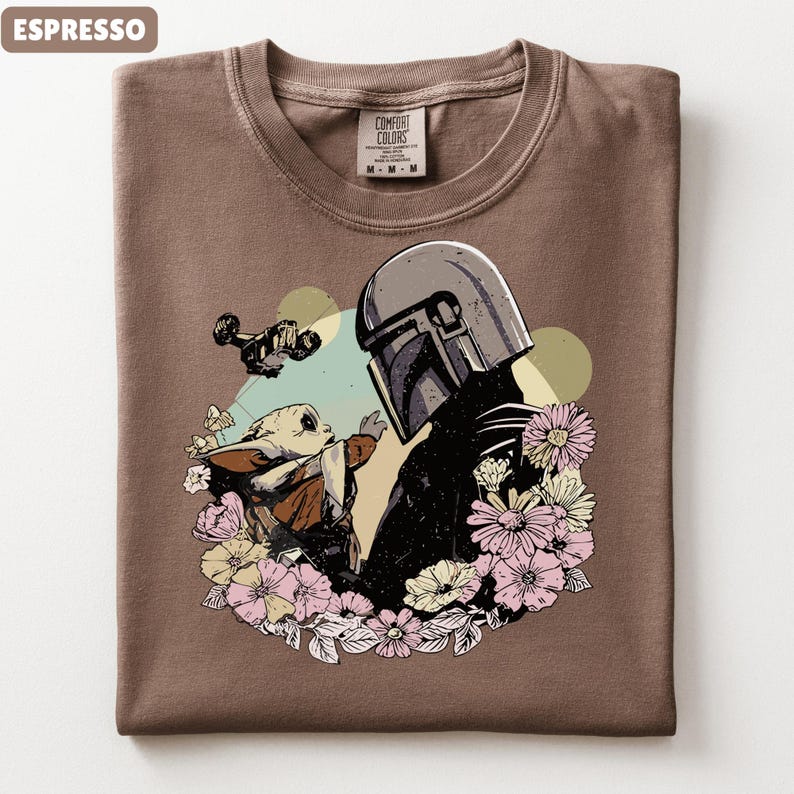 May include: Espresso-colored t-shirt featuring a graphic of a helmeted figure and a small creature surrounded by flowers. The design includes a spaceship and a light green and yellow background. The shirt has a crew neck and a Comfort Colors tag.