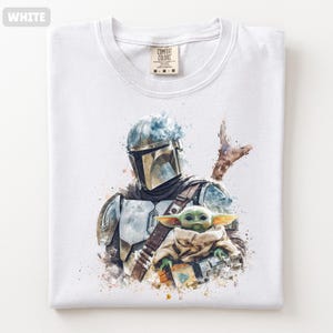 Puede incluir: Camiseta blanca con una ilustraci&oacute;n en acuarela de un personaje mandaloriano y un peque&ntilde;o alien&iacute;gena verde. El dise&ntilde;o incluye armadura, un casco y un bl&aacute;ster. La camiseta est&aacute; doblada y tiene una etiqueta Comfort Colors.