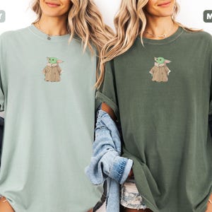 May include: Two t-shirts, one in bay green and the other in moss green, each featuring an embroidered image of a character. The character is green with a tan coat. The shirts have rolled-up sleeves and are worn with denim jackets.