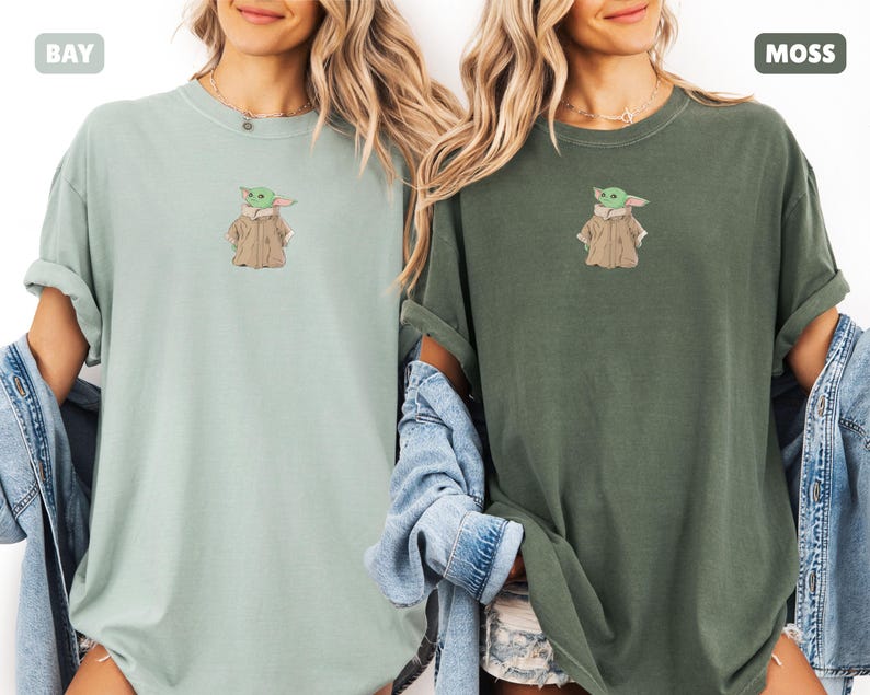 May include: Two t-shirts, one in bay green and the other in moss green, each featuring an embroidered image of a character. The character is green with a tan coat. The shirts have rolled-up sleeves and are worn with denim jackets.