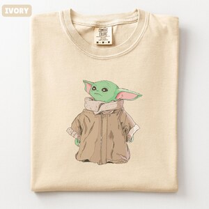 May include: Ivory-colored t-shirt featuring a cartoon illustration of a green alien character wearing a brown coat. The shirt has a crew neck and a tag that reads "COMFORT COLORS". The shirt is folded.