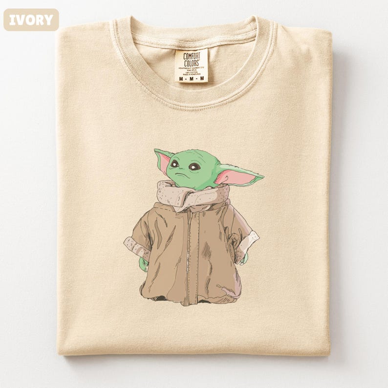 May include: Ivory-colored t-shirt featuring a cartoon illustration of a green alien character wearing a brown coat. The shirt has a crew neck and a tag that reads "COMFORT COLORS". The shirt is folded.