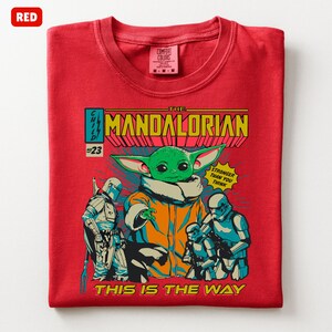 May include: Red t-shirt featuring a comic book style graphic of The Mandalorian. The graphic includes the Child, Stormtroopers, and the text "The Mandalorian", "Stronger Than You Think", and "This Is The Way". The shirt is a Comfort Colors brand.